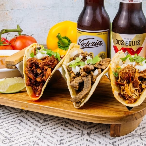 Three tacos with various fillings, lime, salsa, tomatoes, peppers, and two beer bottles.