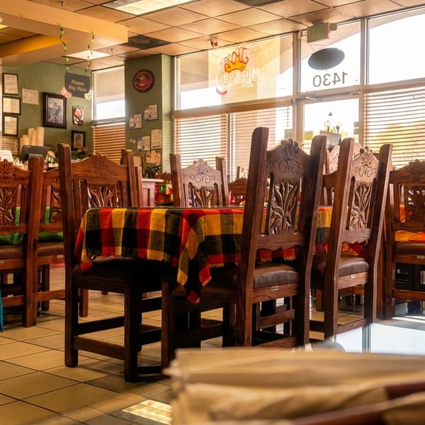 Empty restaurant dining room with carved wooden chairs, colorful tablecloths, and a bar in the background.