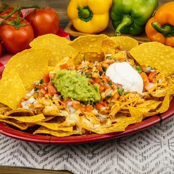 A plate of nachos with guacamole, sour cream, and pico de gallo, surrounded by fresh vegetables.