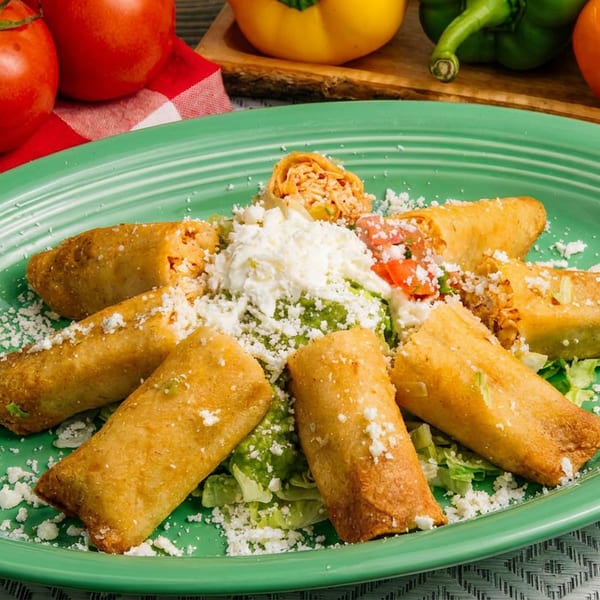 Crispy taquitos on a green plate, garnished with shredded lettuce, guacamole, sour cream, and cheese.