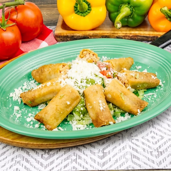 A green plate of golden taquitos with lettuce, cheese, and pico de gallo, surrounded by fresh vegetables.
