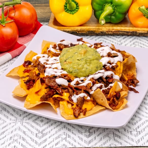 Steak nachos with melted cheese, sour cream, and guacamole, surrounded by fresh tomatoes and bell peppers.